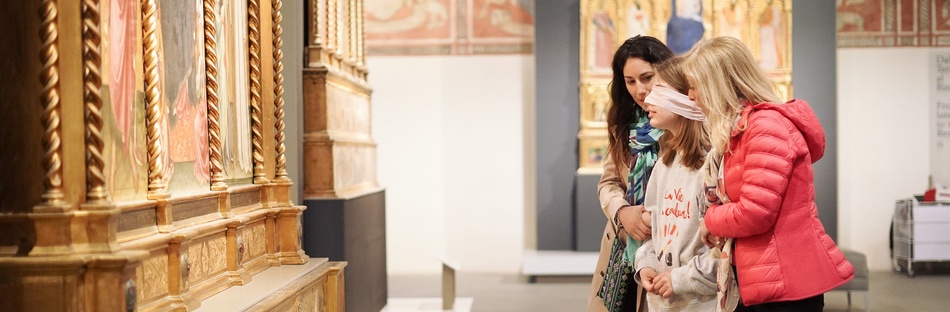 A blindfolded girl supported by two women in front of a work of art